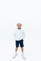 Boy in a white hoodie, navy shorts and white cap standing with his hands in his pockets