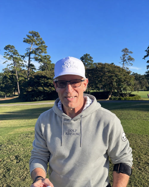 Gray adult Golf Asylum hoodie worn on the course, with the wordmark on the chest, a Ballhead sleeve patch and a smiley-print hood lining